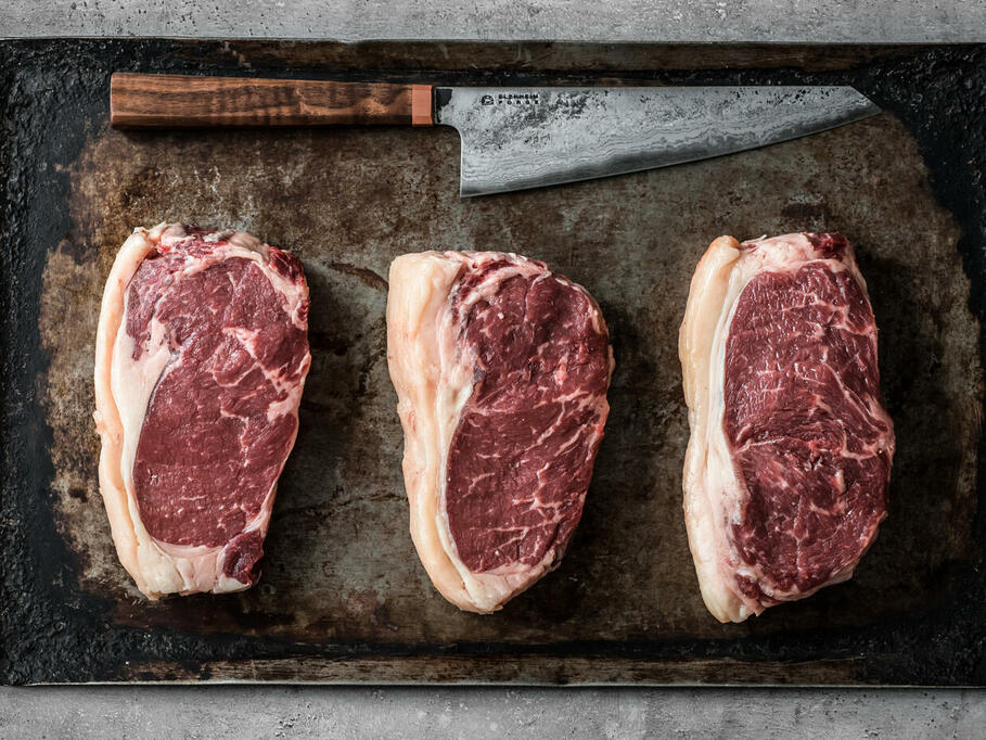 three raw beef steaks lay flat on a baking tray with a sharp knife above them